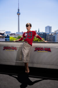 ok se sportska estetika sve više seli izvan terena i postaje deo svakodnevne mode, Zendaya je u Berlinu pokazala kako retro inspiracija može da izgleda savremeno i elegantno. Za fotografisanje povodom filma Spider-Man: Brand New Day, glumica je odabrala kombinaciju koja priziva duh američkog sportskog stila iz sedamdesetih, ali uz prepoznatljivu dozu sofisticiranosti. Iza njenog izdanja stajao je Law Roach, stilista sa kojim Zendaya godinama sarađuje na kreiranju modnih trenutaka koji privlače pažnju. Ovoga puta izbor je pao na komade iz kolekcije kuće Coach za sezonu jesen/zima 2026, koju je dizajnirao Stuart Vevers. Inspiracija za kolekciju pronađena je u američkoj sportskoj kulturi prošlog veka — od univerzitetskih uniformi do klasičnih dresova koji su obeležili jednu eru. Zendayin autfit upravo je preuzeo te elemente: prugasti top nalik sportskom dresu u crveno-roze tonovima, sa istaknutim brojem 39, bio je glavni komad kombinacije. Uz njega je nosila kariranu midi suknju u tamnim nijansama, čime je sportski karakter ublažen ženstvenijim krojem. Iako je isti izgled na pisti bio predstavljen uz patike i visoke sportske čarape, Zendaya ga je prilagodila svom stilu i odlučila se za salonke u čokoladno-braon boji. Detalji su imali važnu ulogu u celokupnom izdanju. Zlatne minđuše, sat Rolex i nakit dali su kombinaciji elegantniji izgled, dok su posebnu pažnju privukli njen dijamantski verenički prsten i zlatna burma koju nosi nakon venčanja sa Tom Hollandom. Holland se pojavio uz Zendayu tokom fotografisanja u Berlinu, a za razliku od njenog promišljeno stilizovanog autfita, izabrao je ležerniji izgled. Svetloplava karirana košulja, farmerke, kožna jakna u boji peska i crne čizme činili su jednostavnu kombinaciju koja se uklopila u njegov opušteni stil. Par je nedavno potvrdio da su u braku, a njihovo zajedničko pojavljivanje izazvalo je veliku pažnju javnosti, posebno zbog činjenice da se njihovi modni izbori često prirodno dopunjuju. Nakon zvaničnog fotografisanja, Zendaya je promenila izdanje za susret sa fanovima. Umesto sportskog retro izgleda, pojavila se u crnom kožnom kompletu, koji je kombinovala sa upečatljivim plavim minđušama u obliku paukove mreže dizajnerke Lydia Courteille. Iako je promenila garderobu, jedan detalj ostao je isti — prstenje koje je nosila kao deo svog novog životnog poglavlja. Zendayin berlinski izgled uklapa se u širi modni trend u kojem sportski elementi dobijaju potpuno novu ulogu. Nekada rezervisani za terene i slobodno vreme, dresovi, karirani komadi i sportske reference danas postaju deo pažljivo osmišljenih modnih kombinacija.